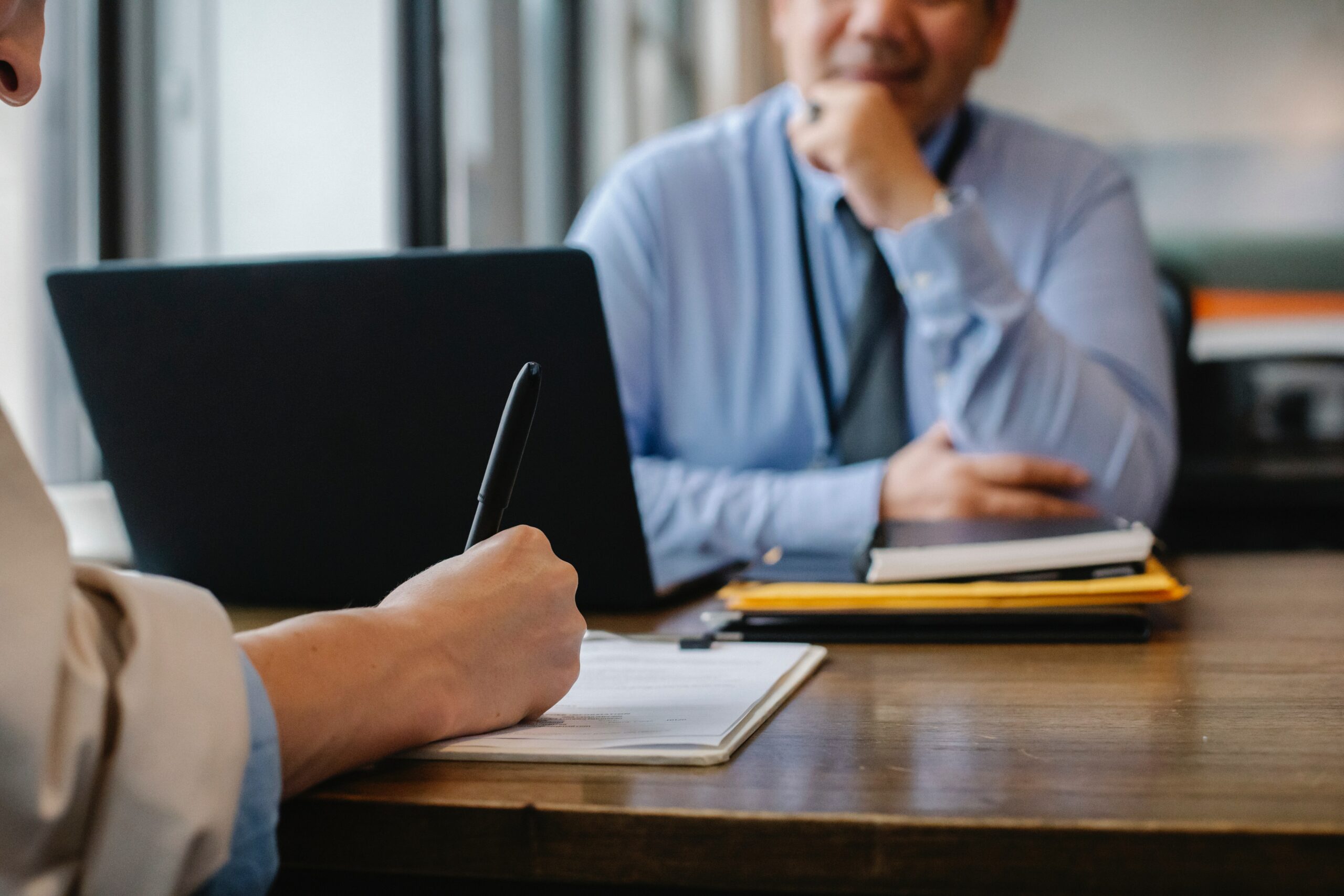 Twee personen tijdens een zakelijk gesprek aan een bureau, waarbij één persoon schrijft op een notitieblok en de ander luistert, met een laptop en documenten op tafel.